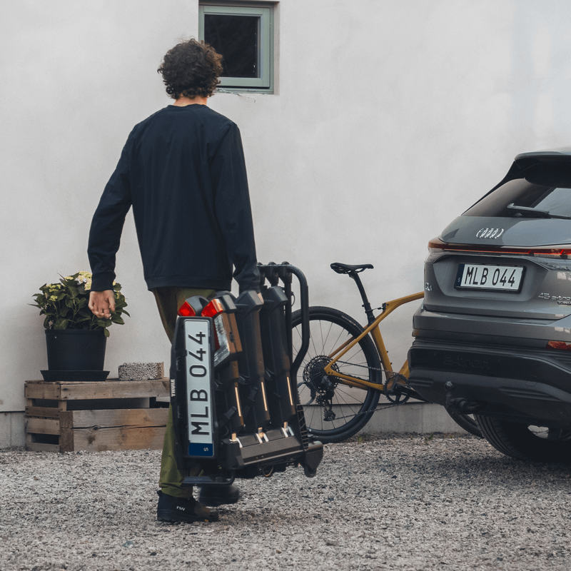 A man in casual outdoor clothing carries a bike rack by its handle, walking between a house and an SUV parked on a gravel driveway.