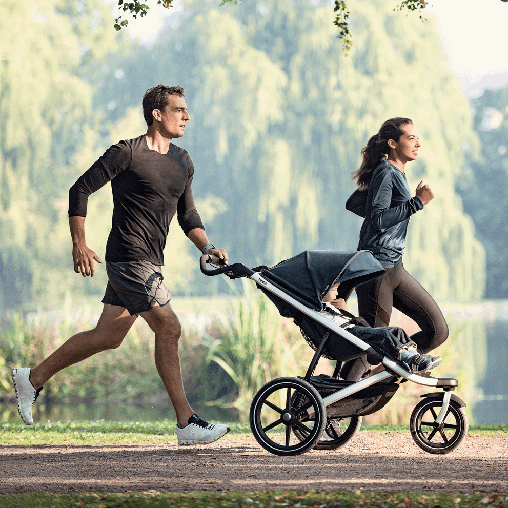 A couple go for a run in a lush green park, pushing a blue Thule Urban Glide 2 jogging stroller.