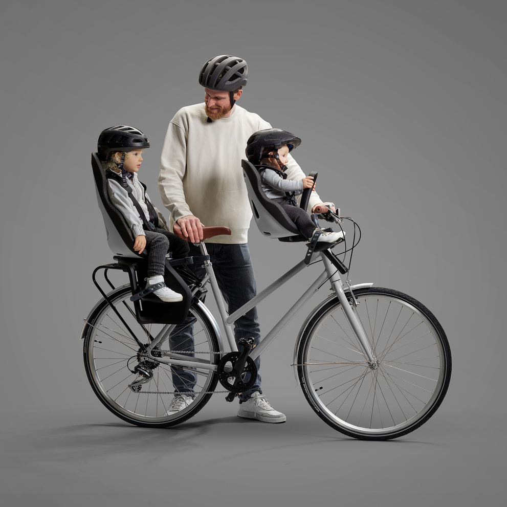 A bearded man stands with a silver bicycle, which has two child bike seats attached: one in front and one at the rear. There is a child in each seat, both wearing black helmets and casual attire