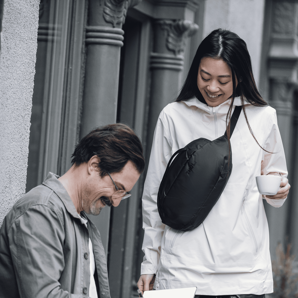 A woman stands outside a cafe carrying a Thule Tact Crossbody bag, talking to a man with a notebook.