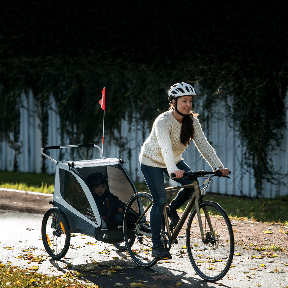 A woman bikes down a bike path with a blue Thule Coaster XT kids' bike trailer.
