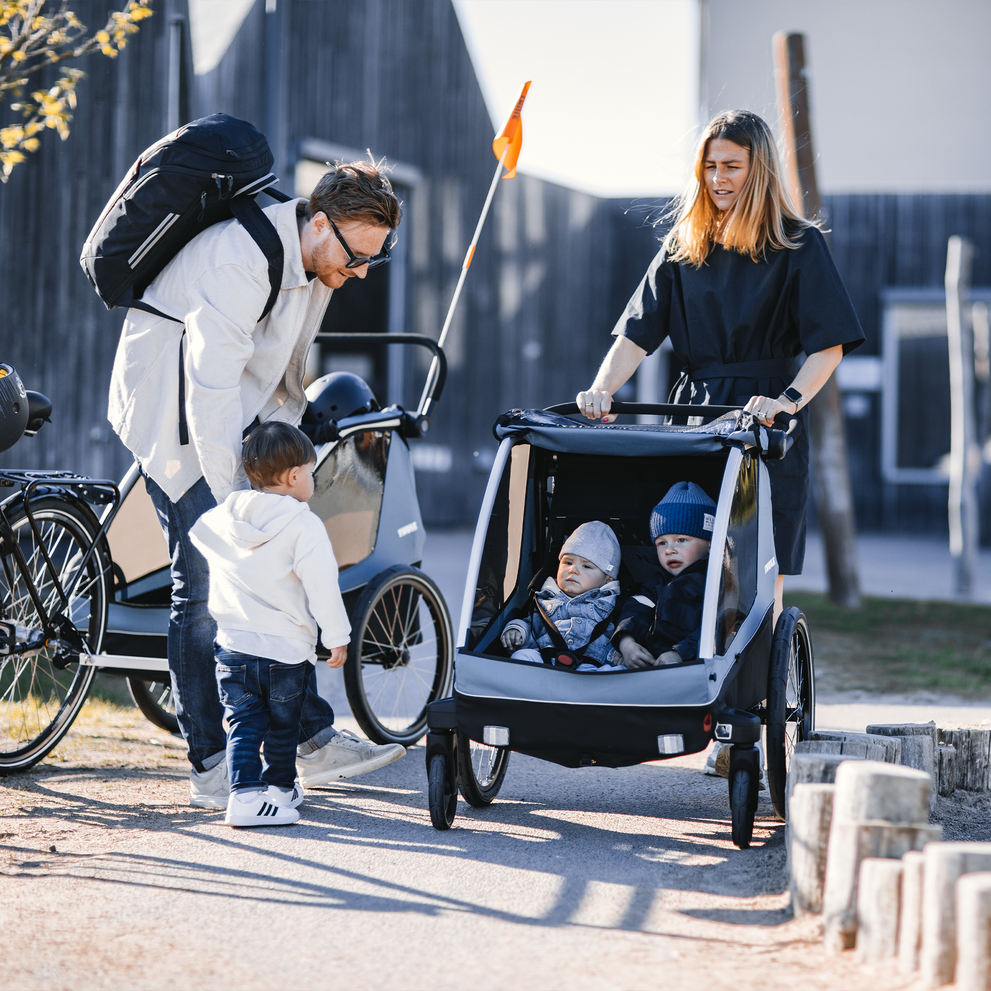 In a park, a father holds his kids' hand and a woman strolls with her kid in a Thule Courier child bike trailer.