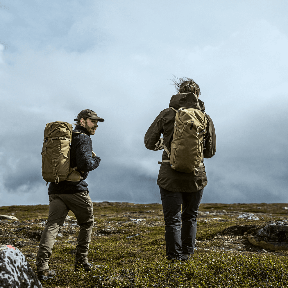 On a cloudy day, two people walk on a field with green Thule AllTrail X 35L hiking backpacks.