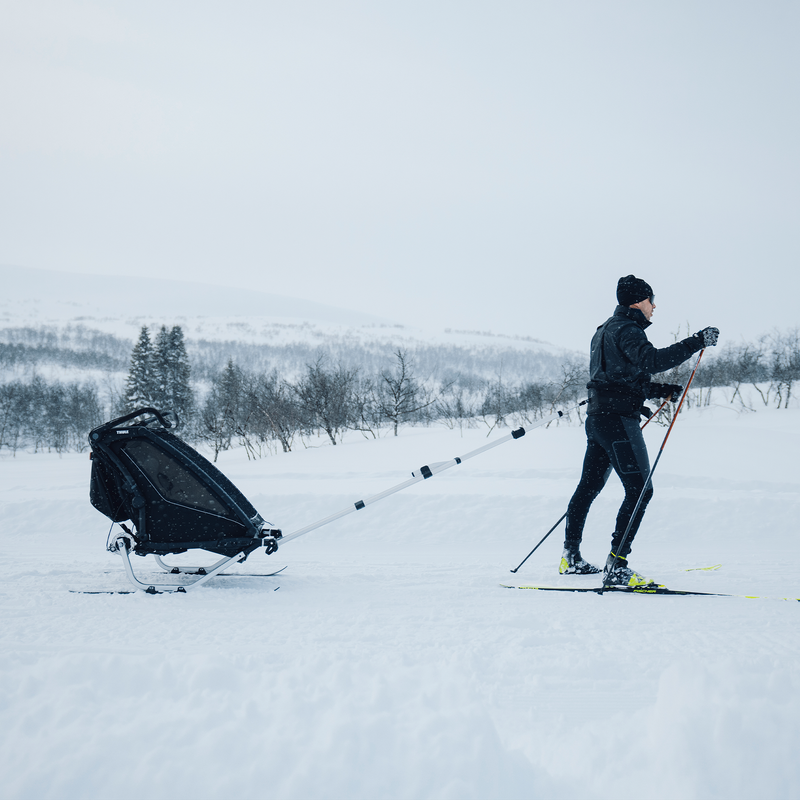 A person skiing in a snowy landscape with a Thule Chariot Sport 2 multisport trailer attached to their body using a ski kit.
