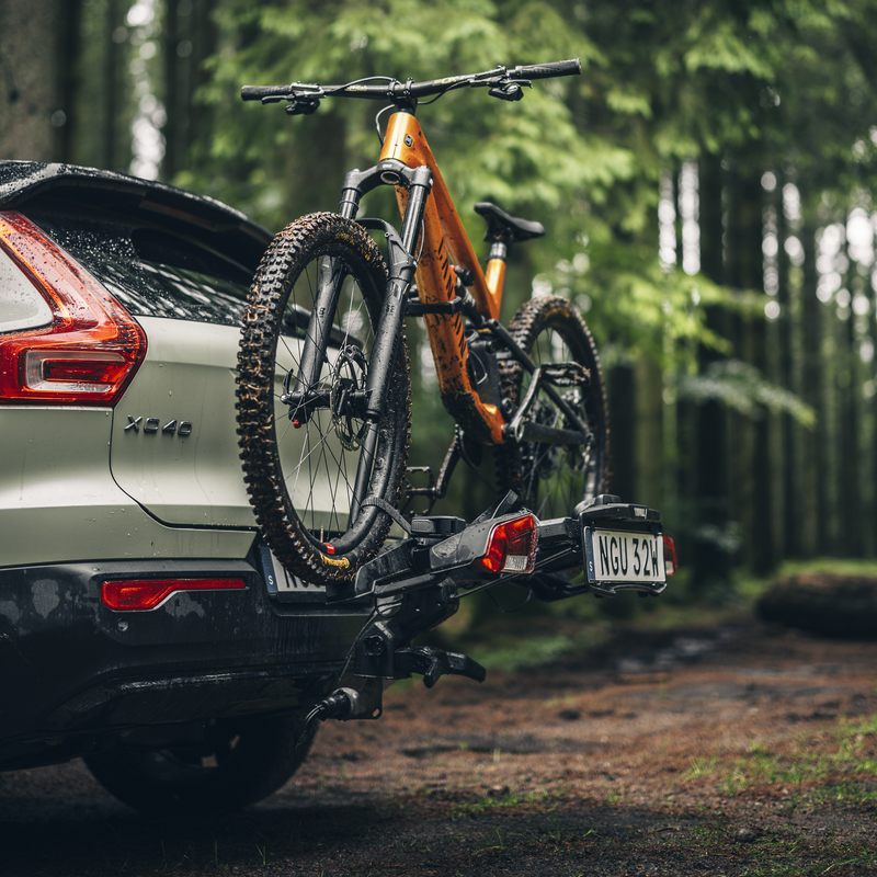 A closeup of a mountain bike on a Thule Outpace towbar bike carrier mounted on a sage green car, parked in the woods.