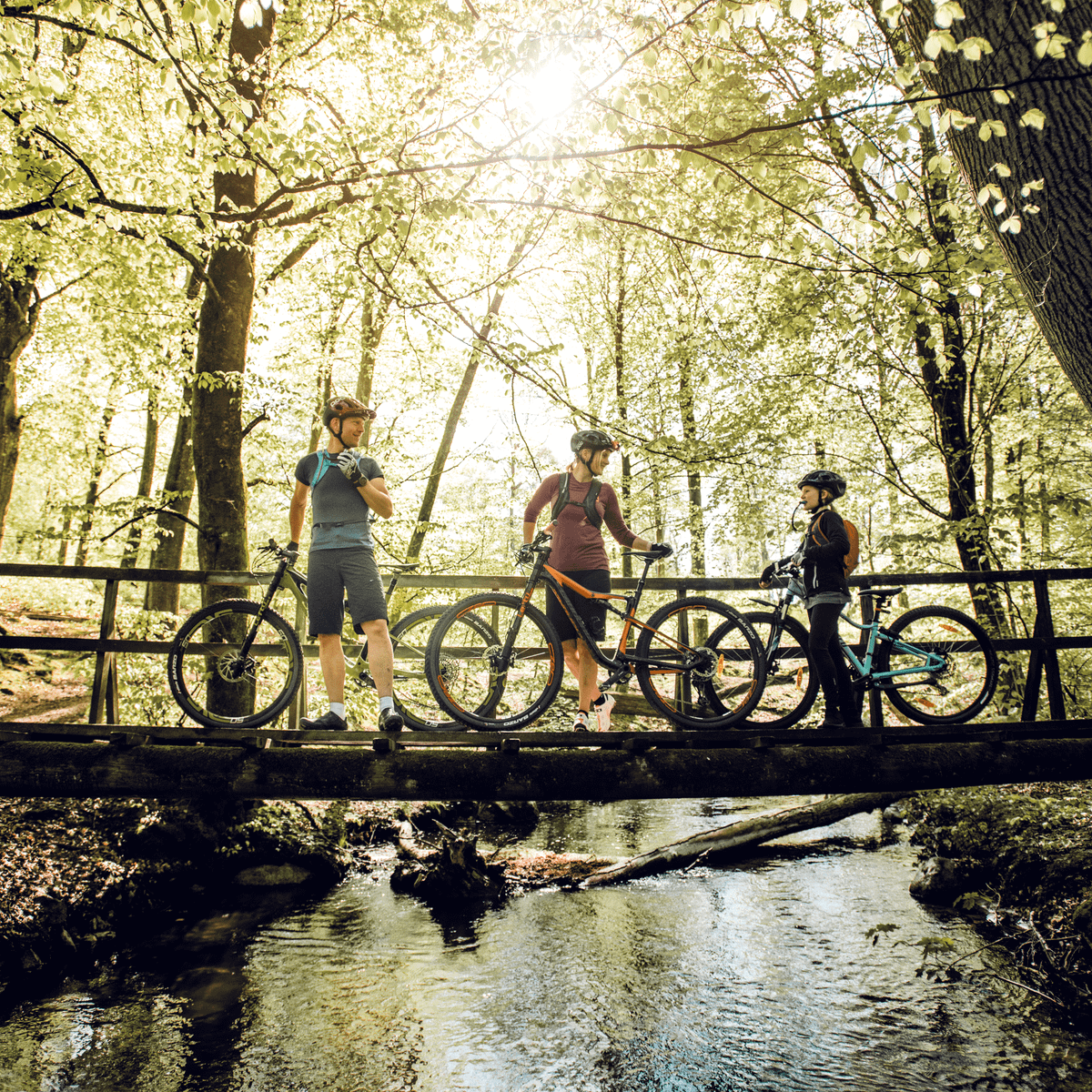 Three cyclists stands on a bridge in the forest on their bikes with Thule UpTake hydration packs.