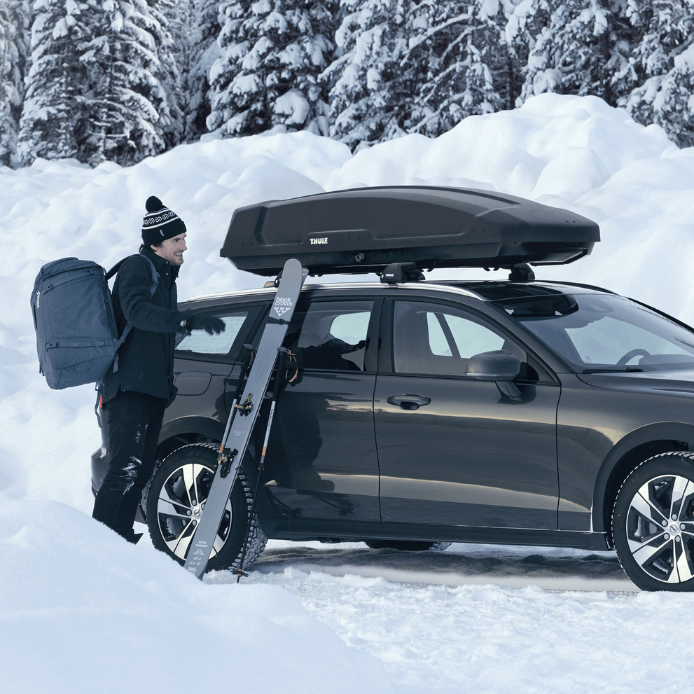 A man unloading the ski gear from a car with a roof top box in a snowy landscape with a cottage in the background