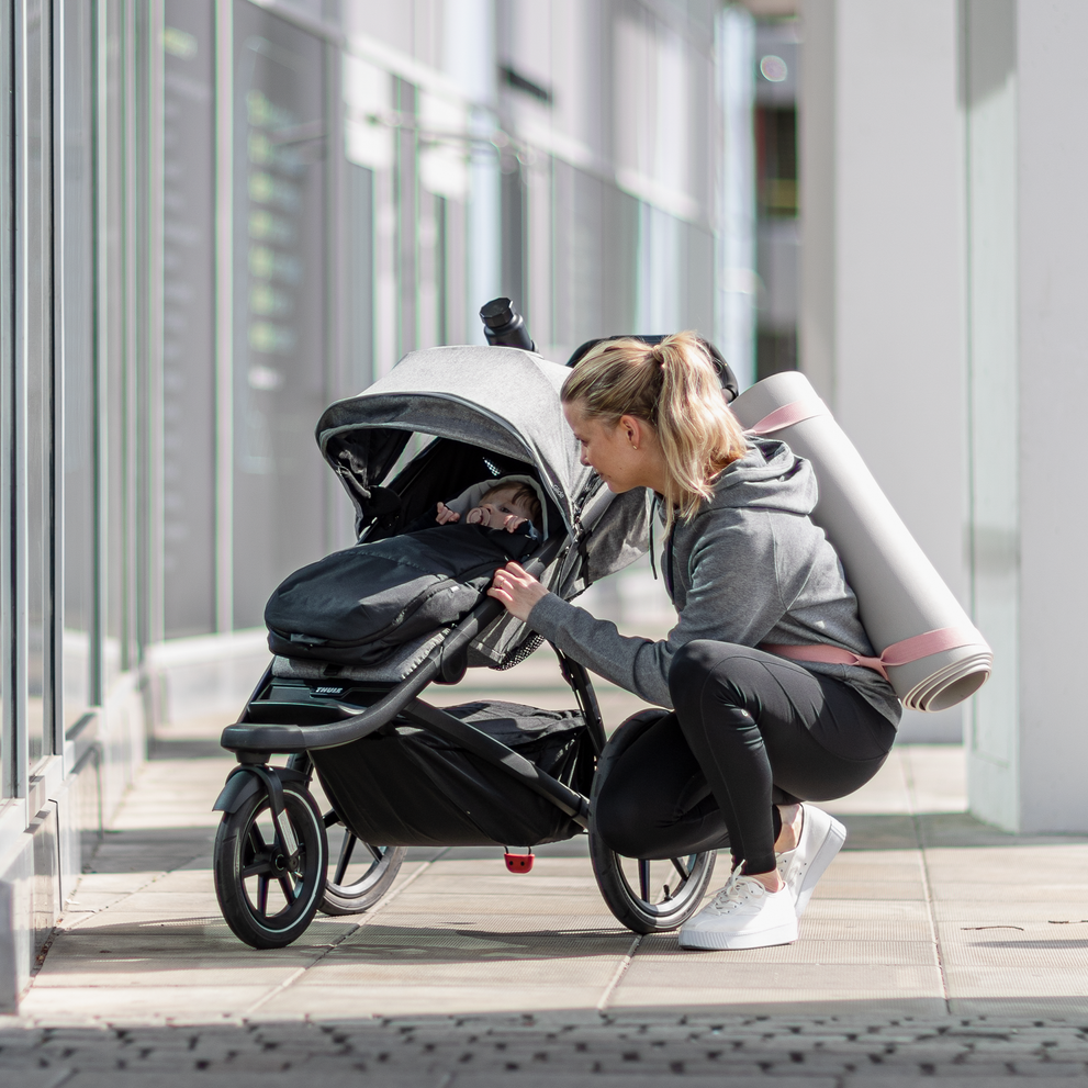 A woman with a yoga mat kneels to speak to her child in a Thule Newborn Nest for Thule strollers.