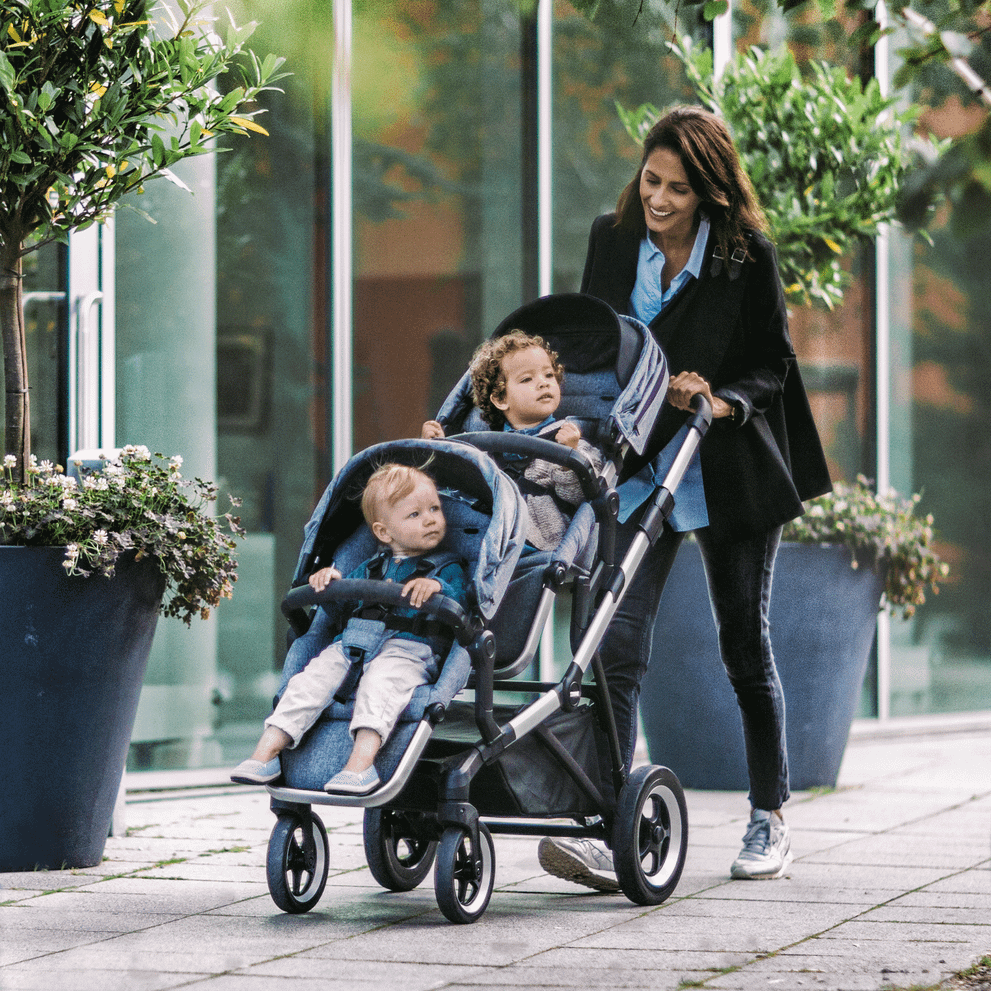 A woman walks down a street with her kids in a stroller that has a blue Thule Sleek Sibling Seat.