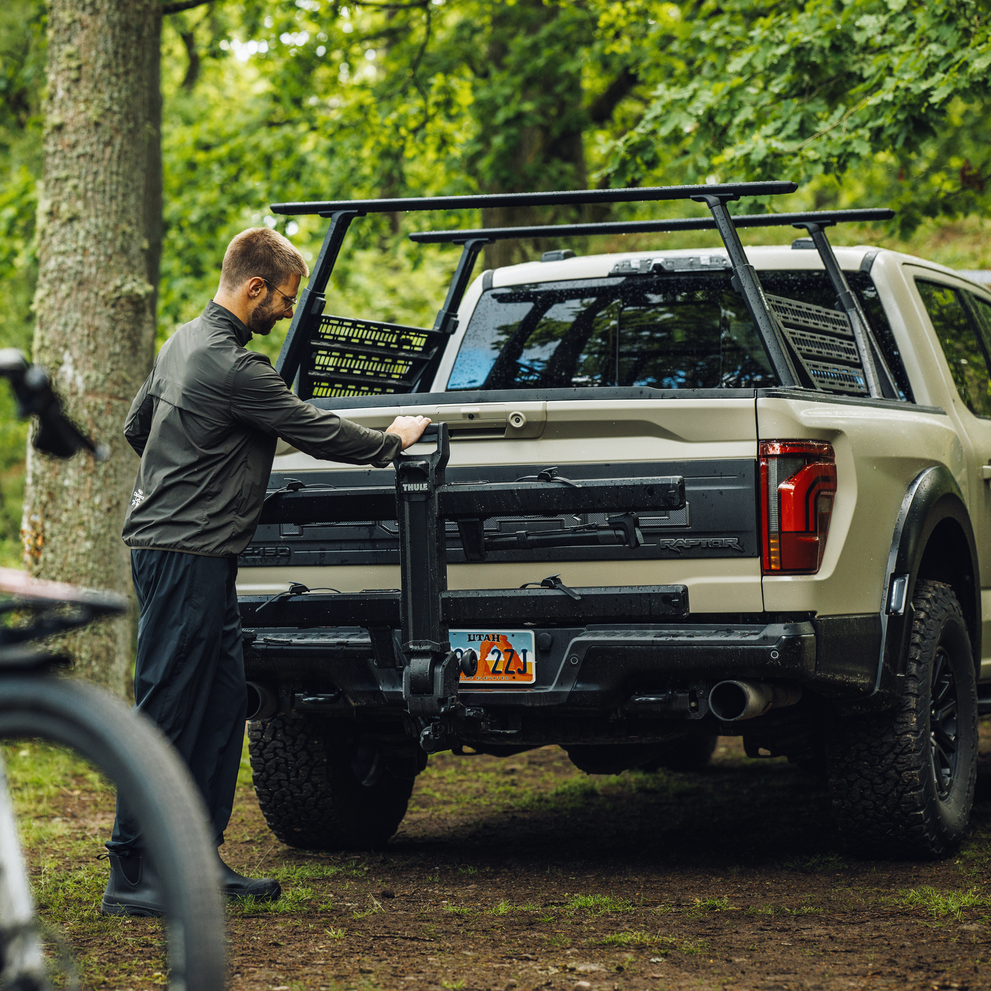 A man folds up the Thule Vero hitch bike rack against the back of a truck, parked in an outdoor setting.