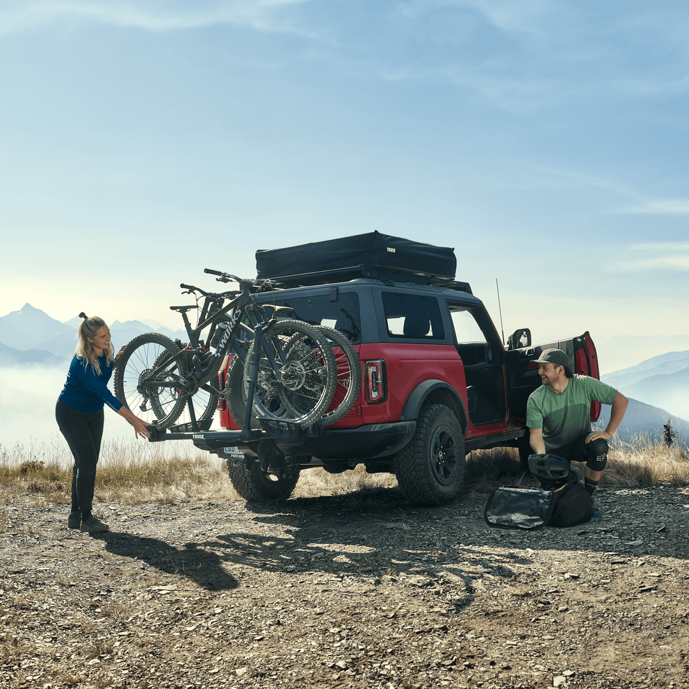 Woman loads bikes on Thule T2 Pro XTR hitch bike rack on a red overlander vehicle in the mountains.