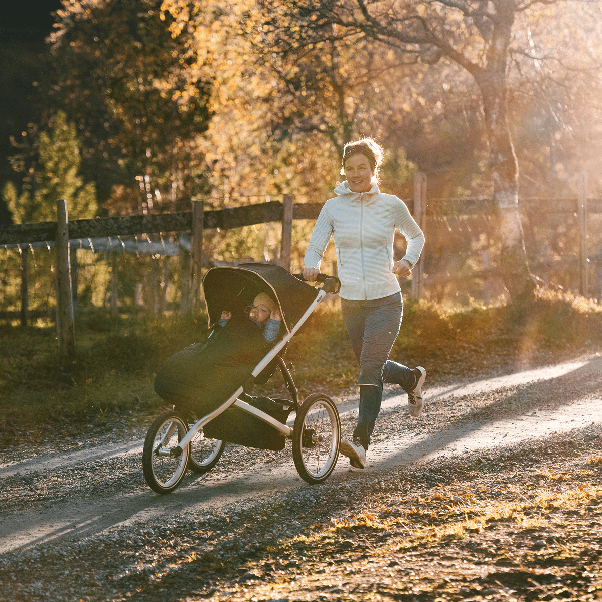A woman jogs on a country path in the sunshine with her child in a black Thule Glide 2 running stroller.