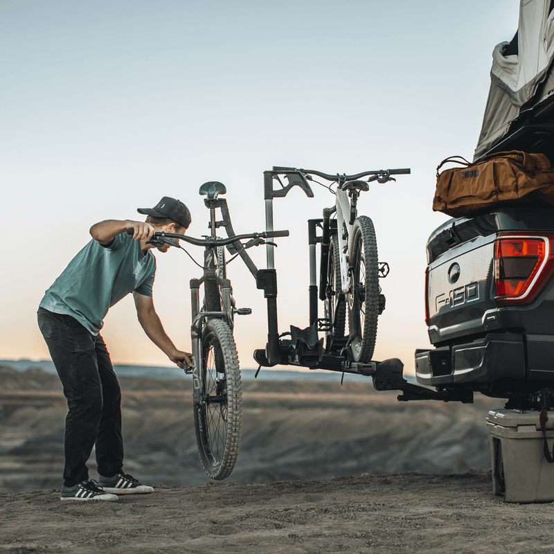 A man loads his bikes onto a Thule Verse bike rack attached to a pick up with a rooftop tent.