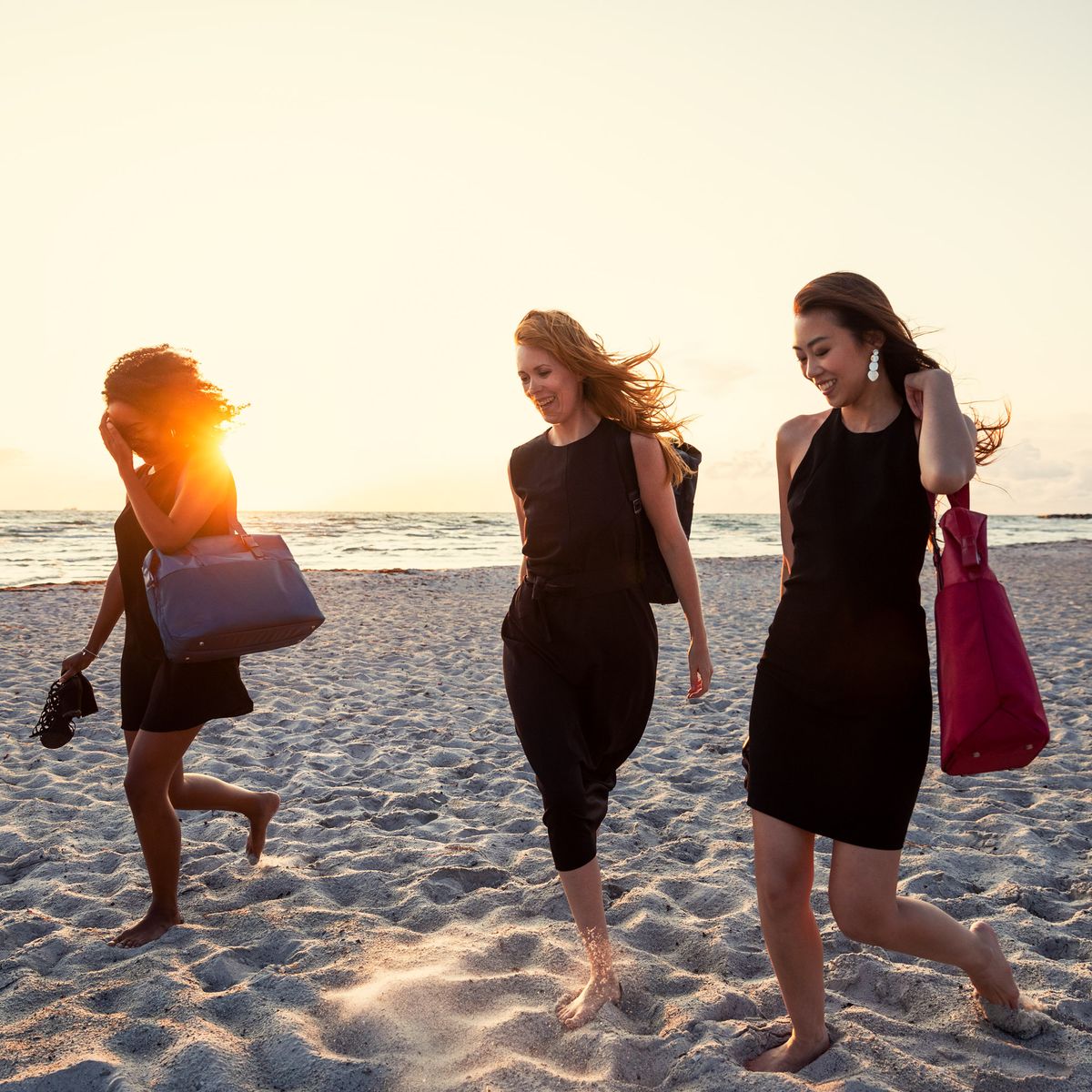 Three women walk down the beach in the sunset carrying Thule Spira backpacks and totes in black and red.