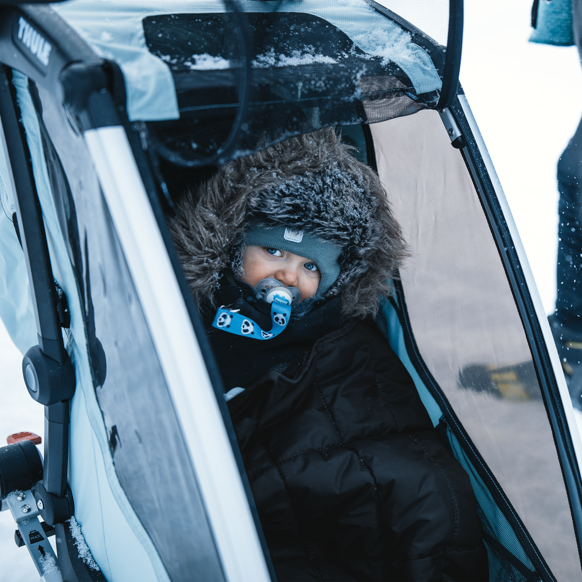 A baby sits in a jogging stroller in the snow with a Thule Footmuff Sport.