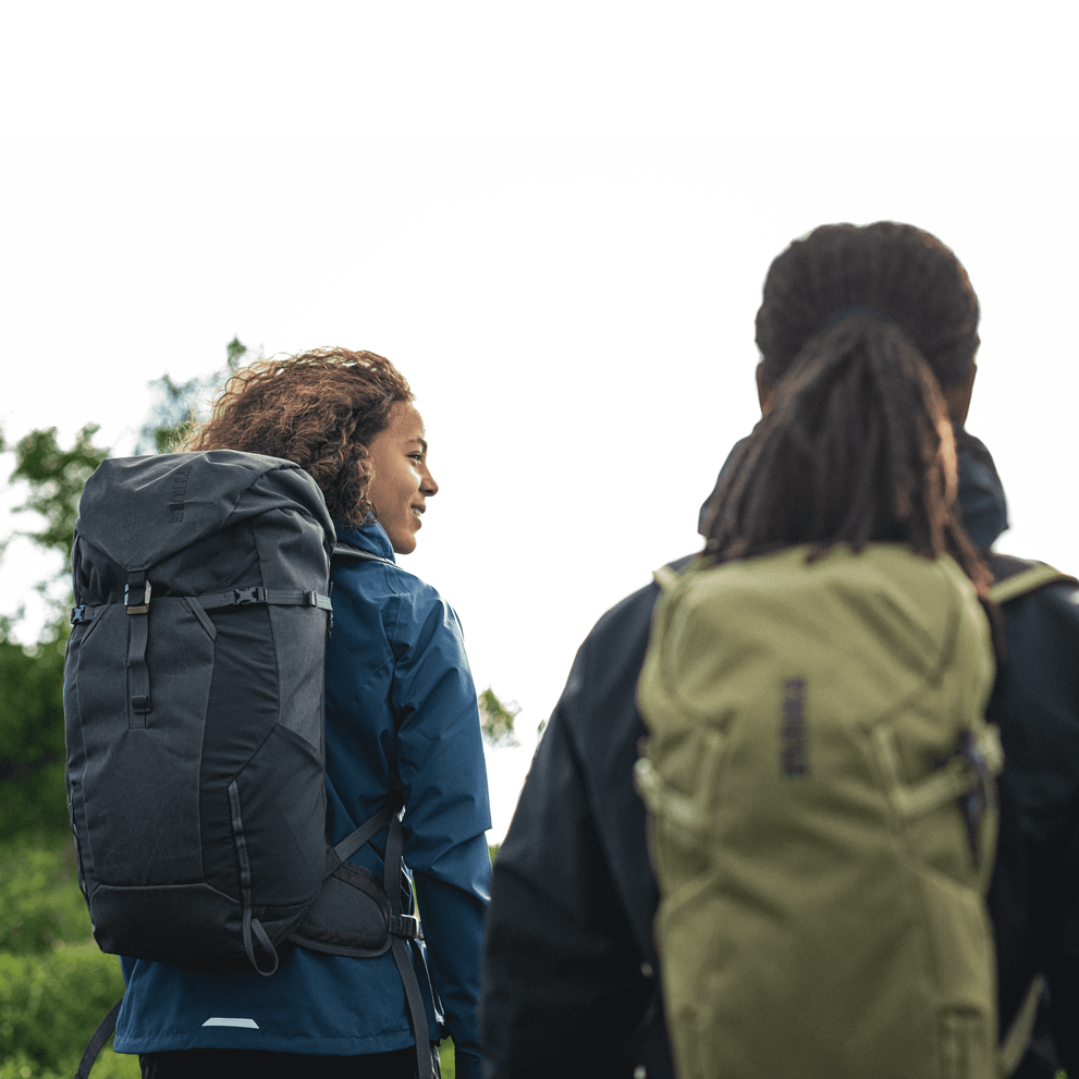 Two people walk on a country path with black and green Thule AllTrail X hiking backpacks.