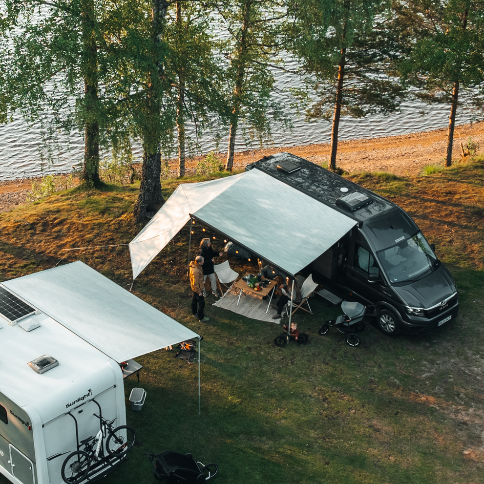 Aerial view of camper vans with Thule awnings near a lakeshore, as people gather under a Thule awning with string lights.