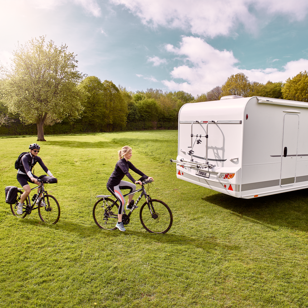 Two cyclists cycle on the grass behind a white motorhome with a Thule Sport G2 Standard rv bike rack.