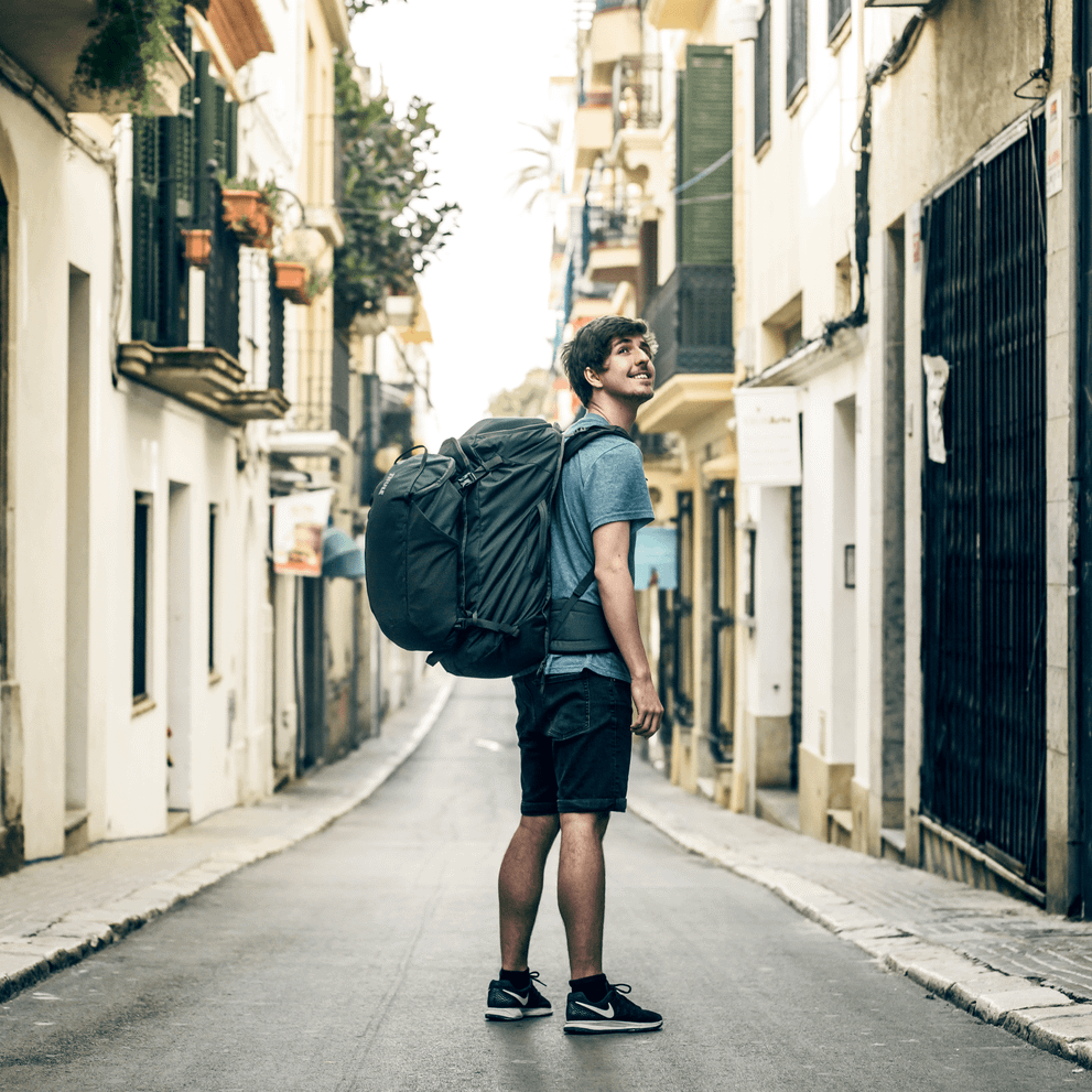 A man stands on an empty street looking up, carrying a black Thule Landmark  70L travel backpack.