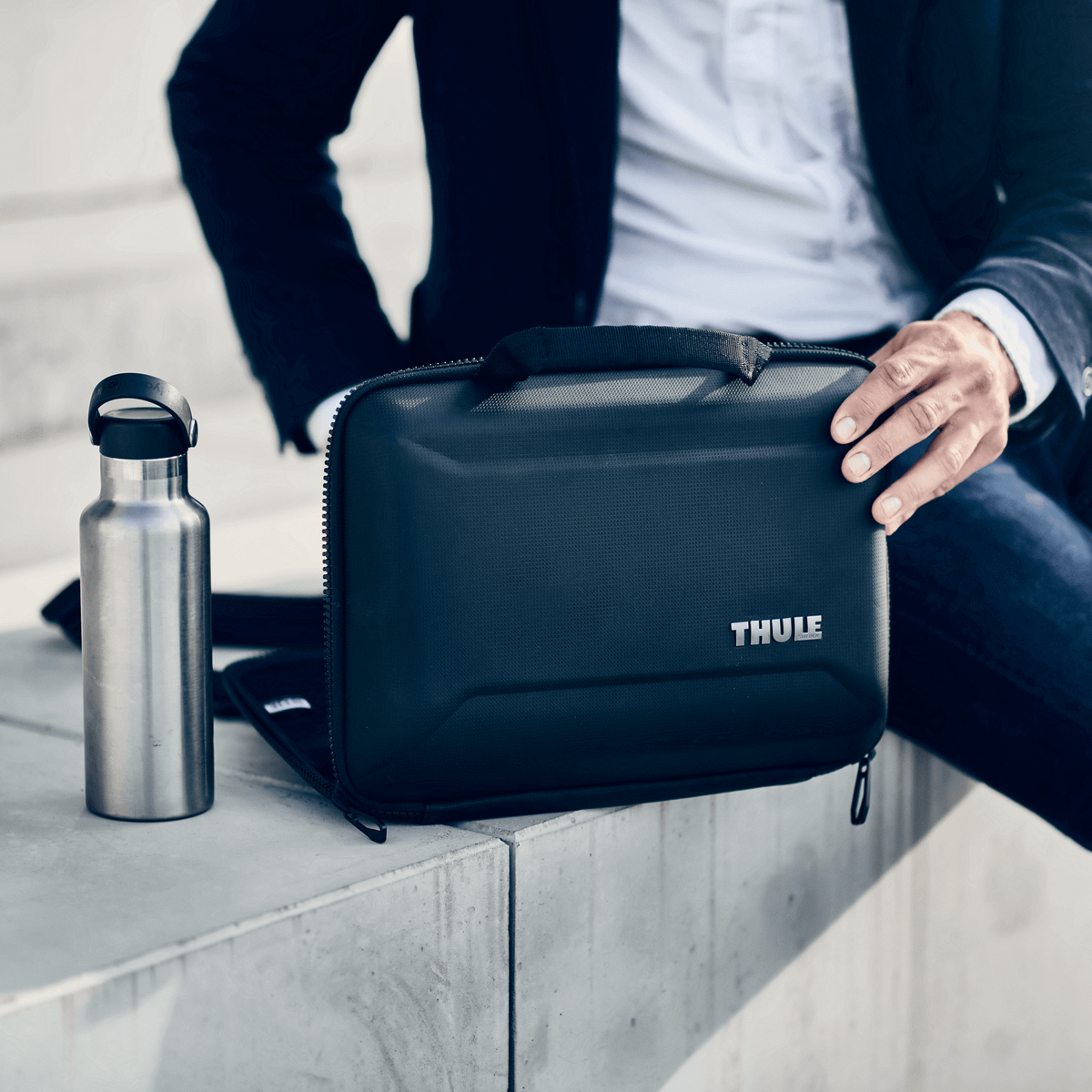 A person wearing professional clothing opens their black Thule Gauntlet 4 MacBook Pro Attaché next to a thermos.