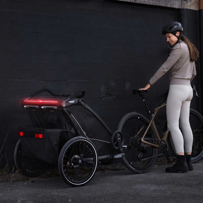 A woman looks back at her black Thule Chariot Sport 2 child bike trailer with integrated LED lights.
