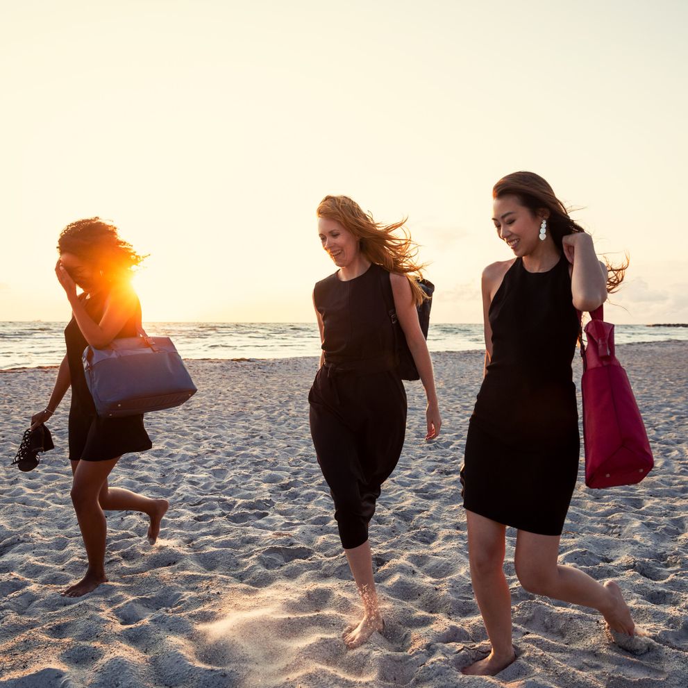 Three women walk down the beach in the sunset carrying Thule Spira backpacks and totes in black and red.