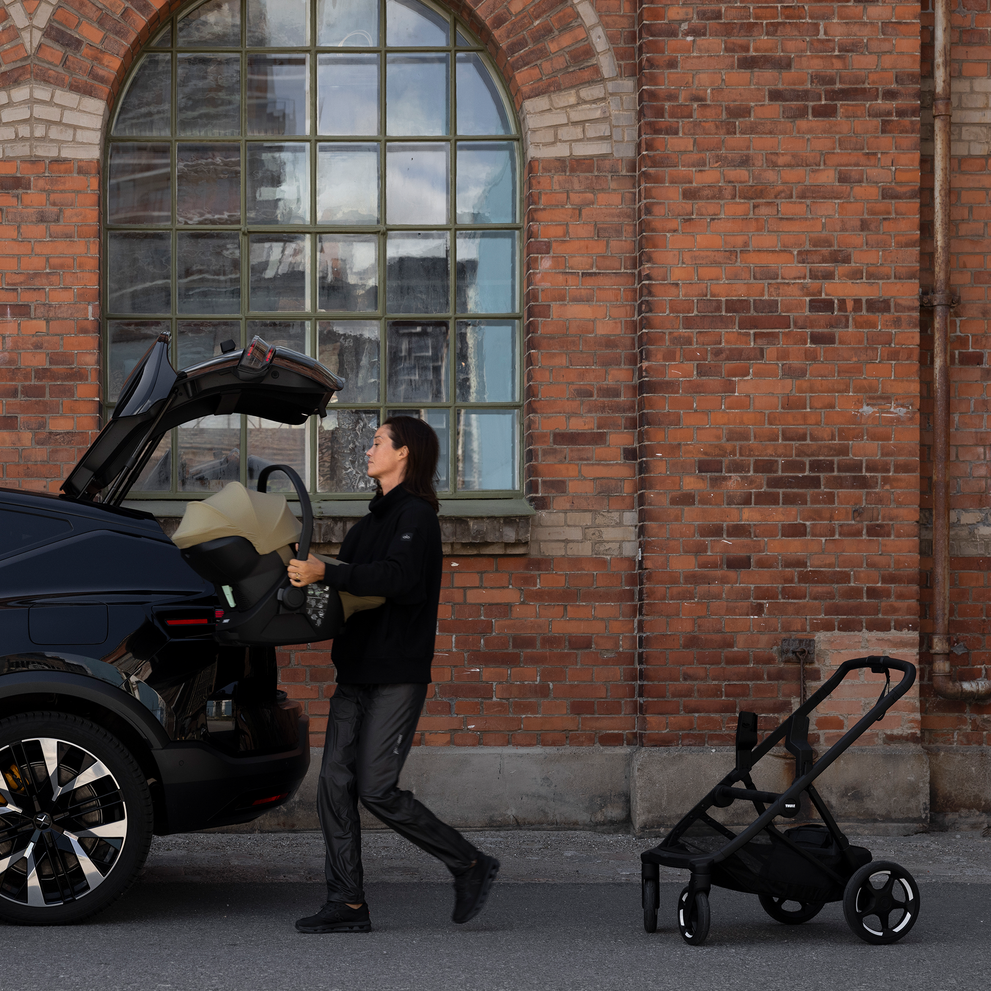 Adult carrying an infant car seat removed from a Thule Charm stroller with car seat adapters beside a vehicle.