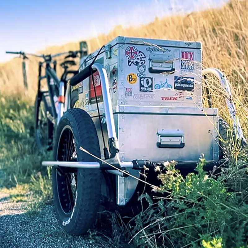 A Reacha City bike trailer attached to a bicycle, parked on a sunny rural road, featuring a storage box covered in stickers.