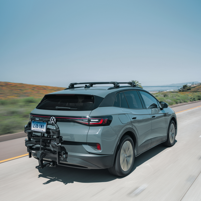 A Volkswagen SUV driving on a coastal road with the Thule OutPace hitch bike carrier in its stowed poisition, equipped with the foldable Thule OutPace license plate holder kit.