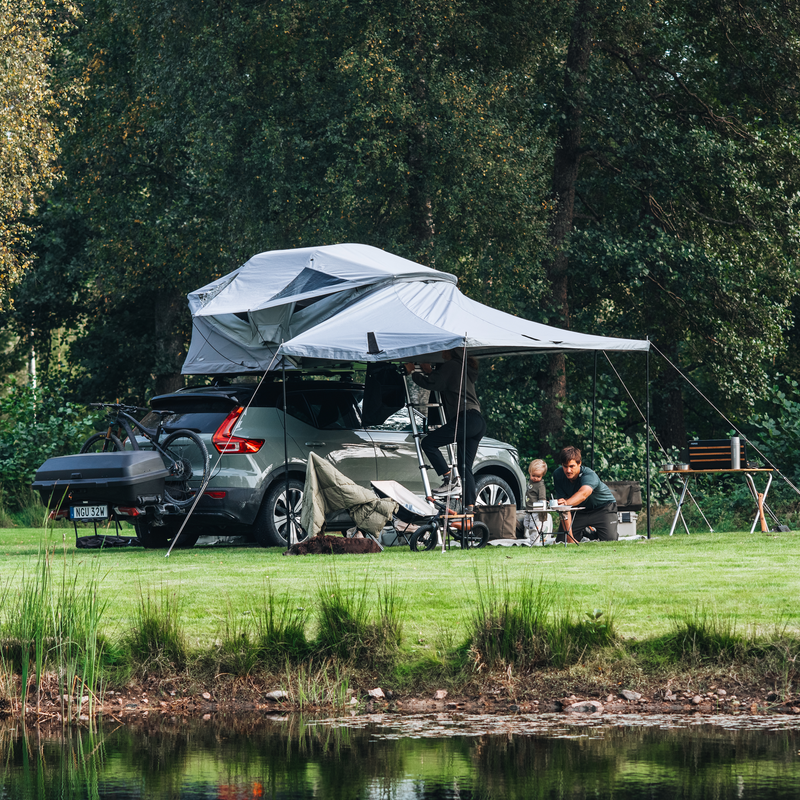Thule Approach 2 soft-shell rooftop tent unfolded and mounted with awning on the top of a car with one bike and Thule Santu cargo box on the towbar bike rack, parked in an outdoor setting and a family interacting by the car.