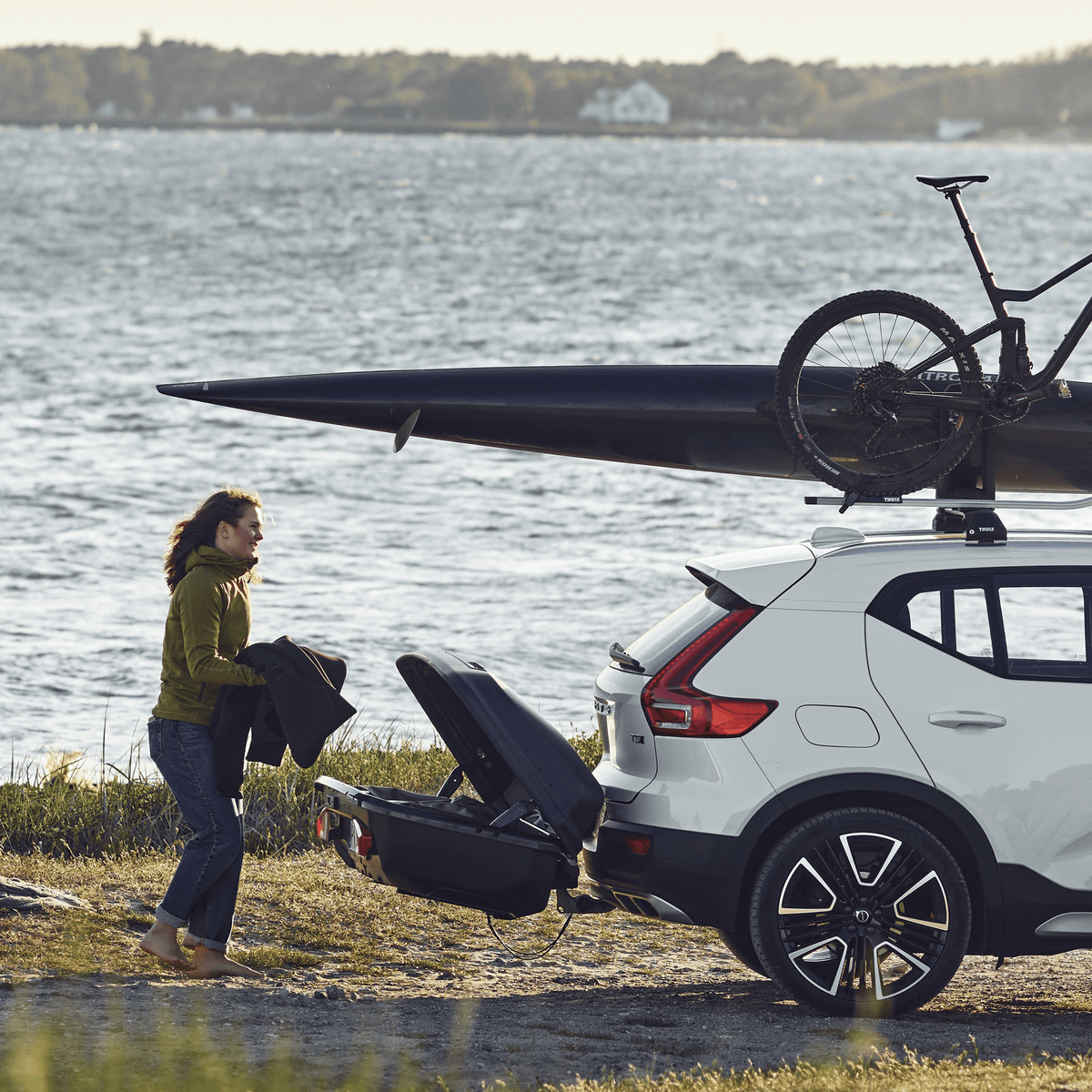 A woman unloading sports gear from a car with a Thule Arcos cargo box and a kayak and a bike mounted on the roof.