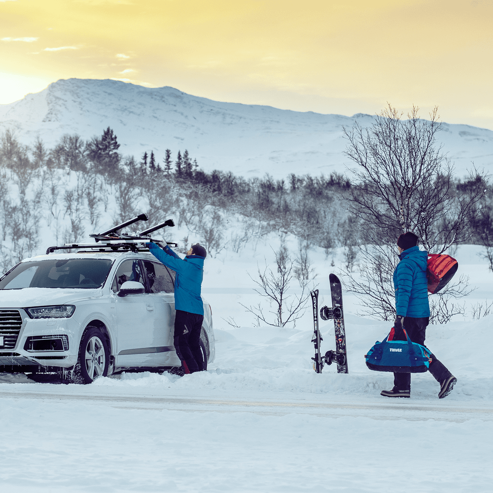 A man is unloading skis from his car roof with a Thule SnowPack Extender while another man is walking towards him with Thule Chasm bags.