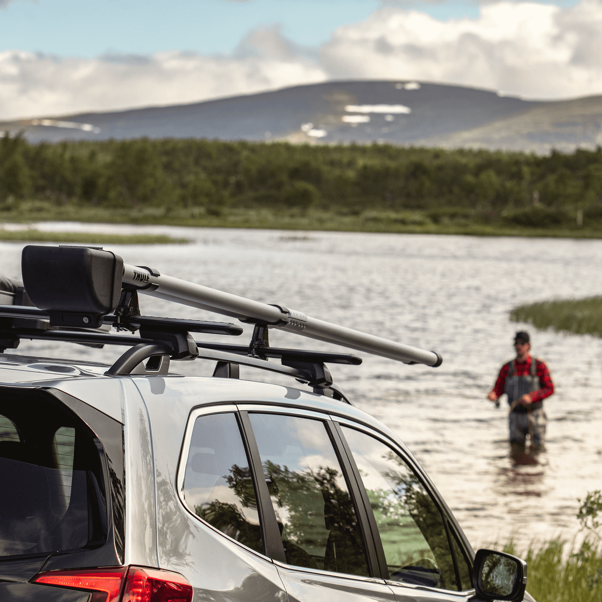 Closeup of a car with a Thule RodVault close to a lake with a man fishing in the background