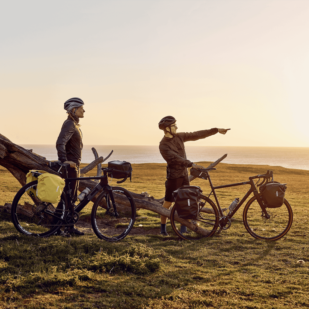 Two cyclists point at the setting sun and ocean with bike carrying 13L Thule Shield bike bags.