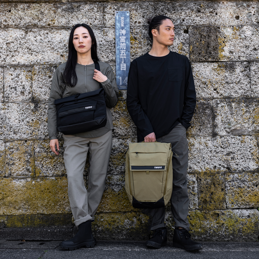 Man and woman stand against a mossy wall with a Thule Paramount 14L messenger bag.