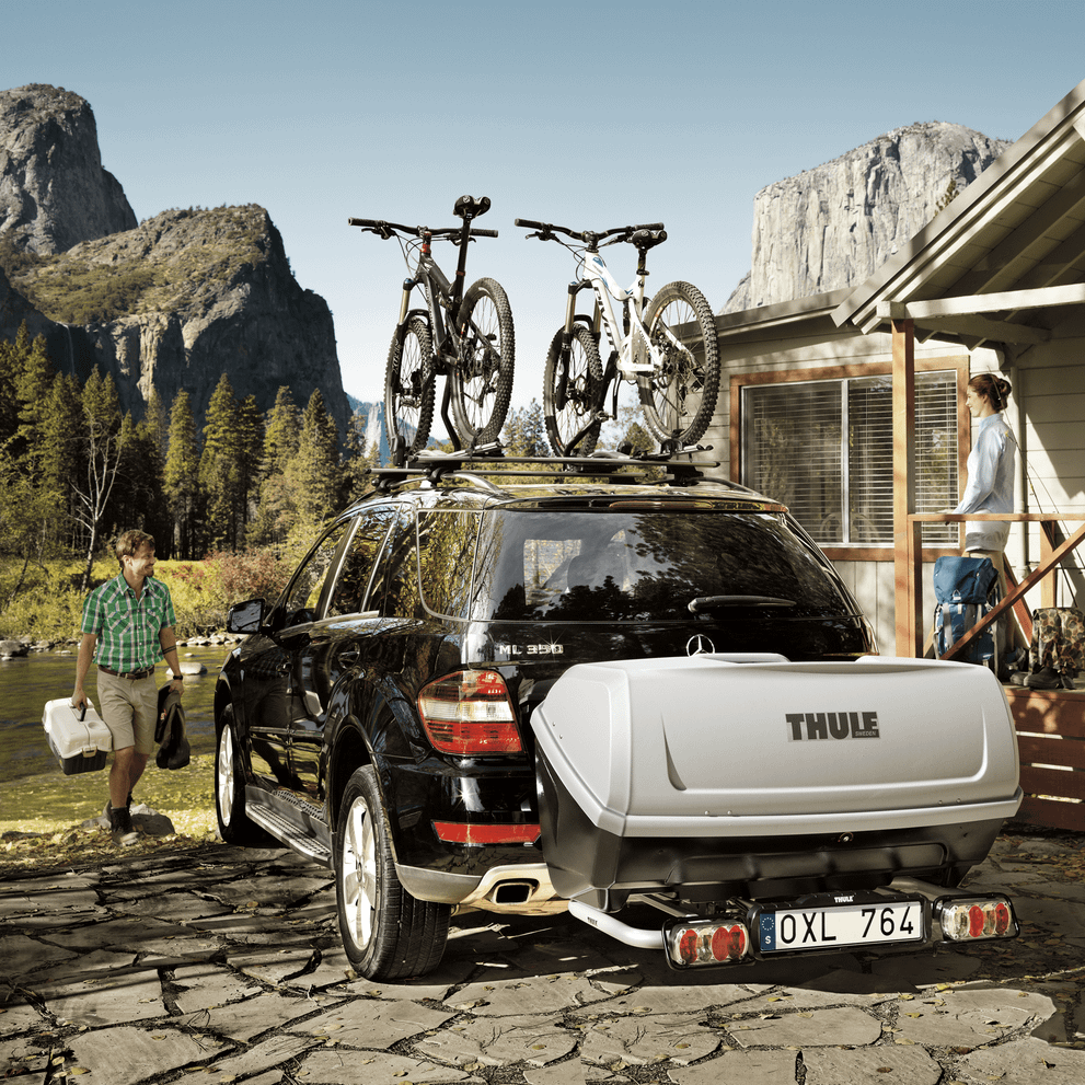 A man is walking towards a car with two bikes on the roof and a Thule BackUp and a Thule Easybase mounted on the back.