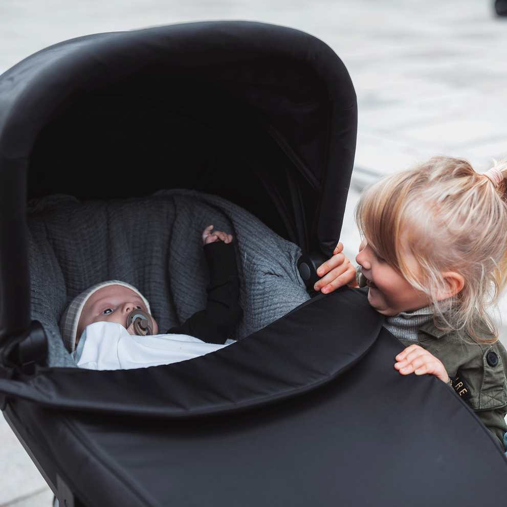 Two toddlers look at a baby lying in the black Thule Urban Glide 2 all-terrain baby stroller.