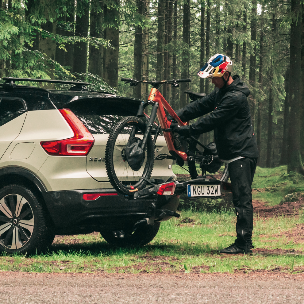 A man is placing a mountain bike on a Thule Outpace towbar bike carrier mounted on a sage green car, parked in the woods.