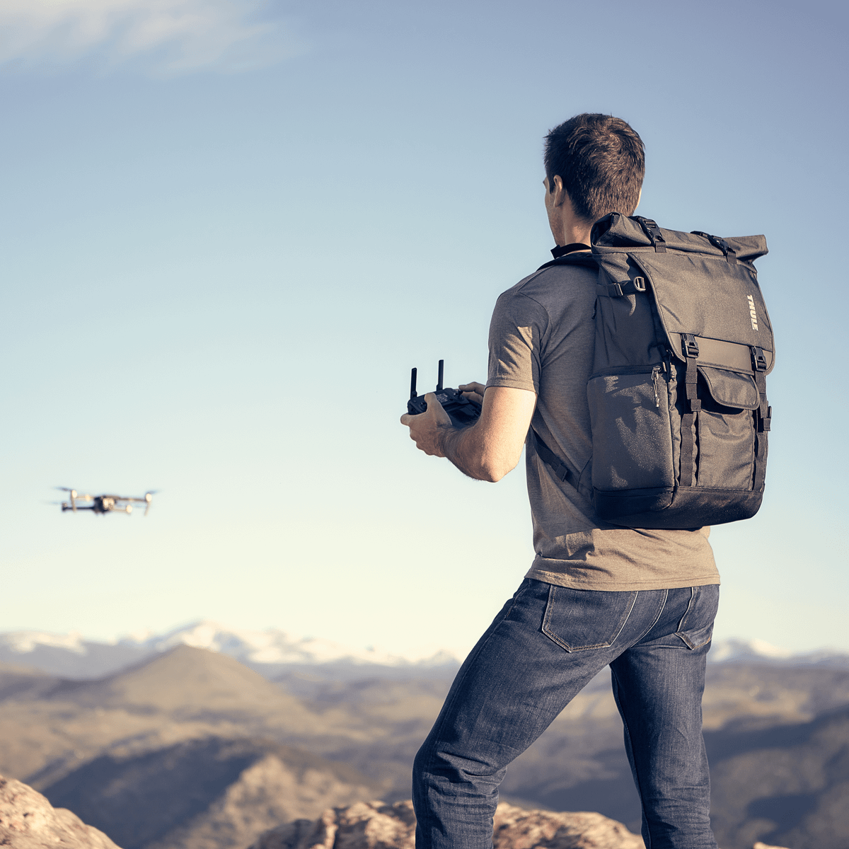 A man stands by mountains controlling a drone, carrying  a Thule Covert DSLR Rolltop backpack.
