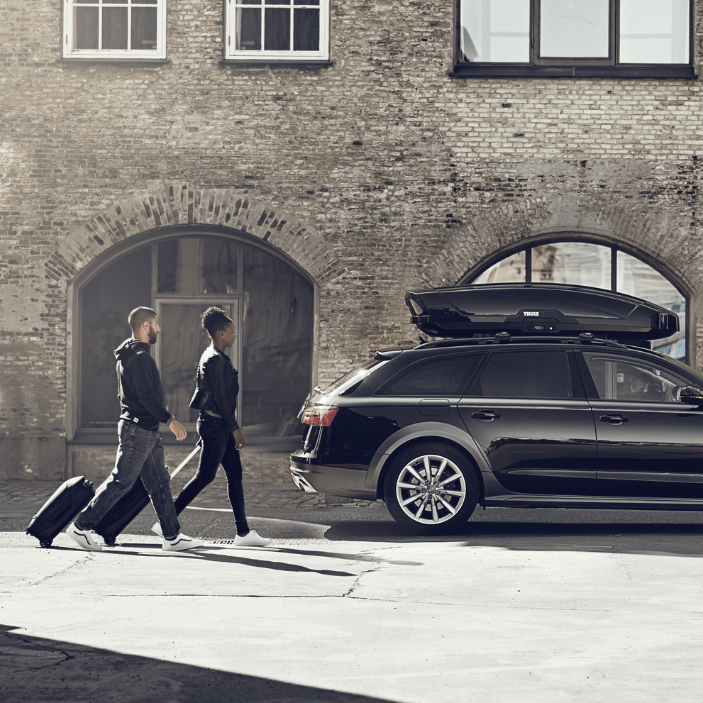 Two men with rolling luggage walking past a parked car with a roof box.