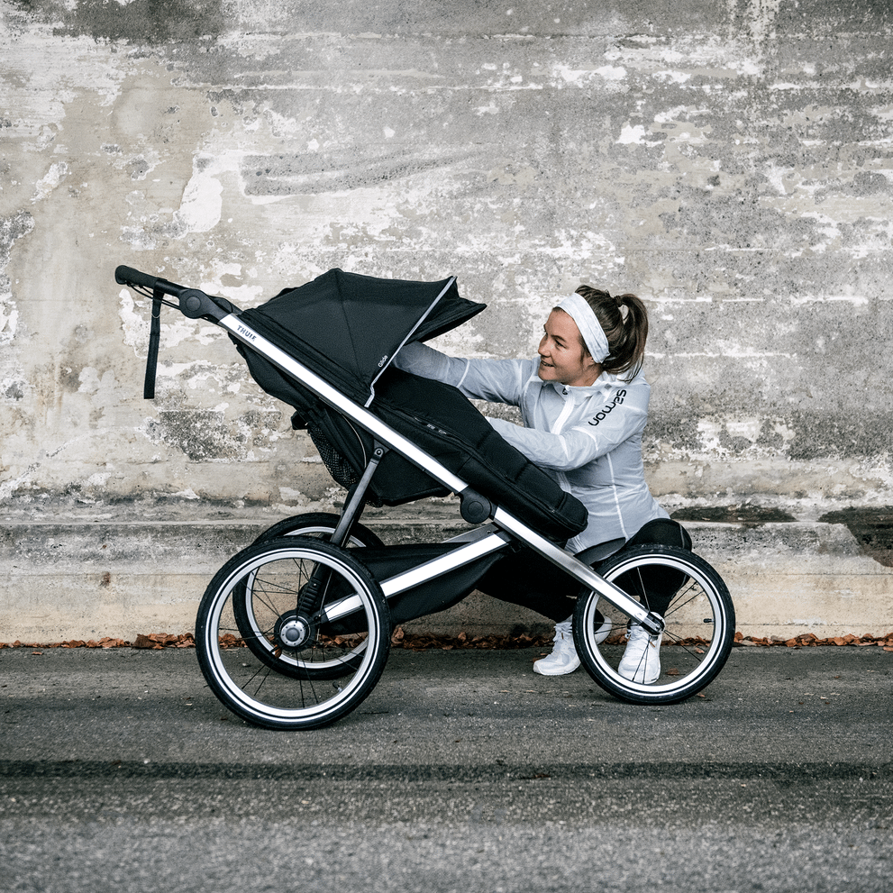 Next to a concrete wall, a woman tends to her child inside a black Thule Glide 2 jogging stroller.