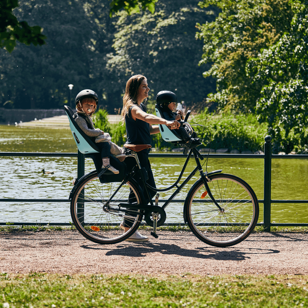 A woman walks in a park with her bike and two children in the rear and front Thule Yepp Nexxt child bike seat.