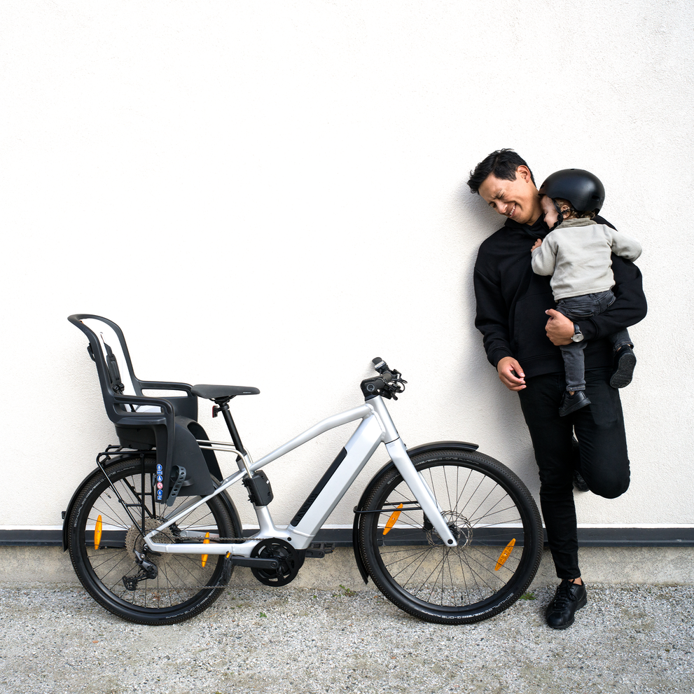 A man leans against a wall with his child and a bike with a Thule RideAlong2 child bike seat.