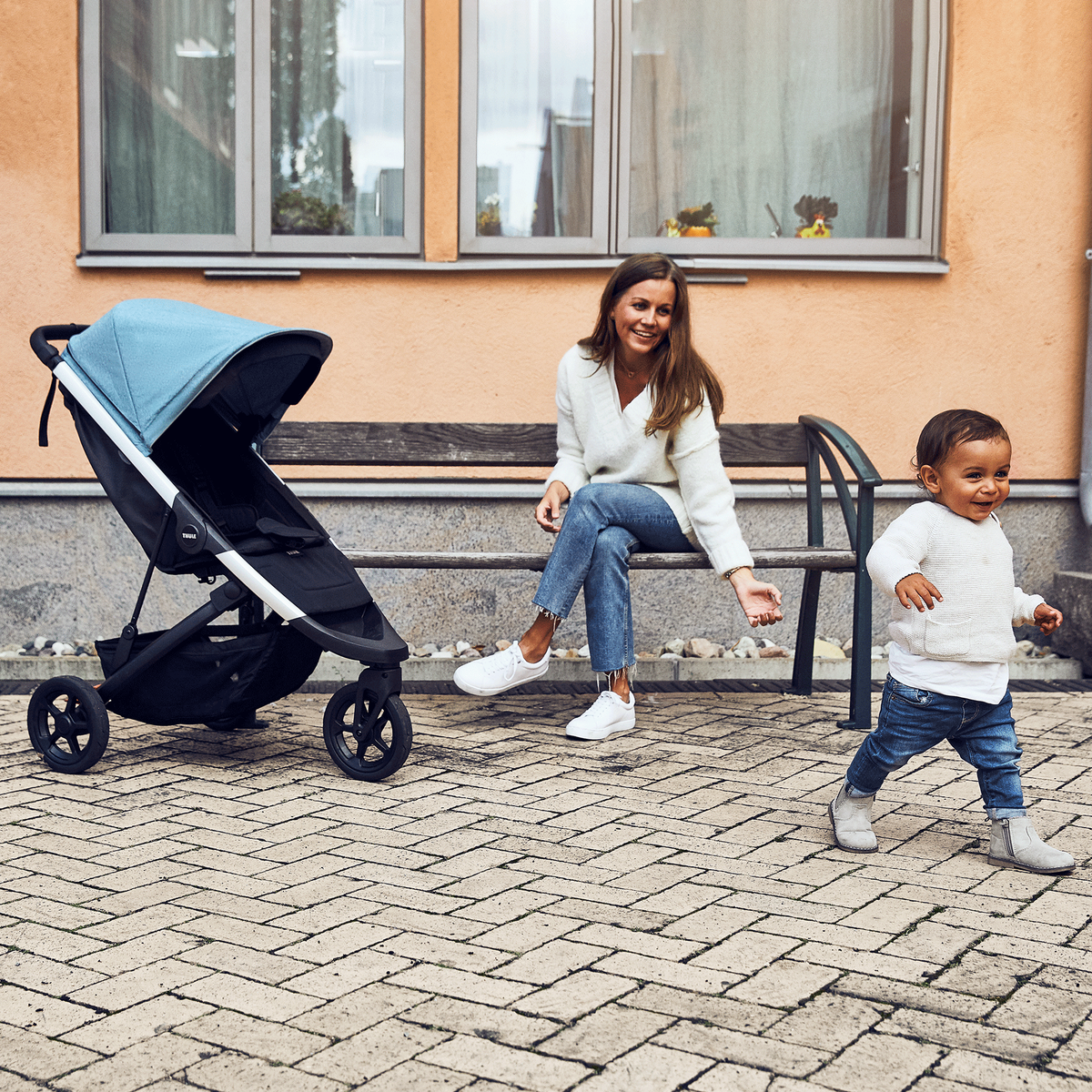 A woman sits on a bench while her child runs around a blue Thule Spring compact stroller.