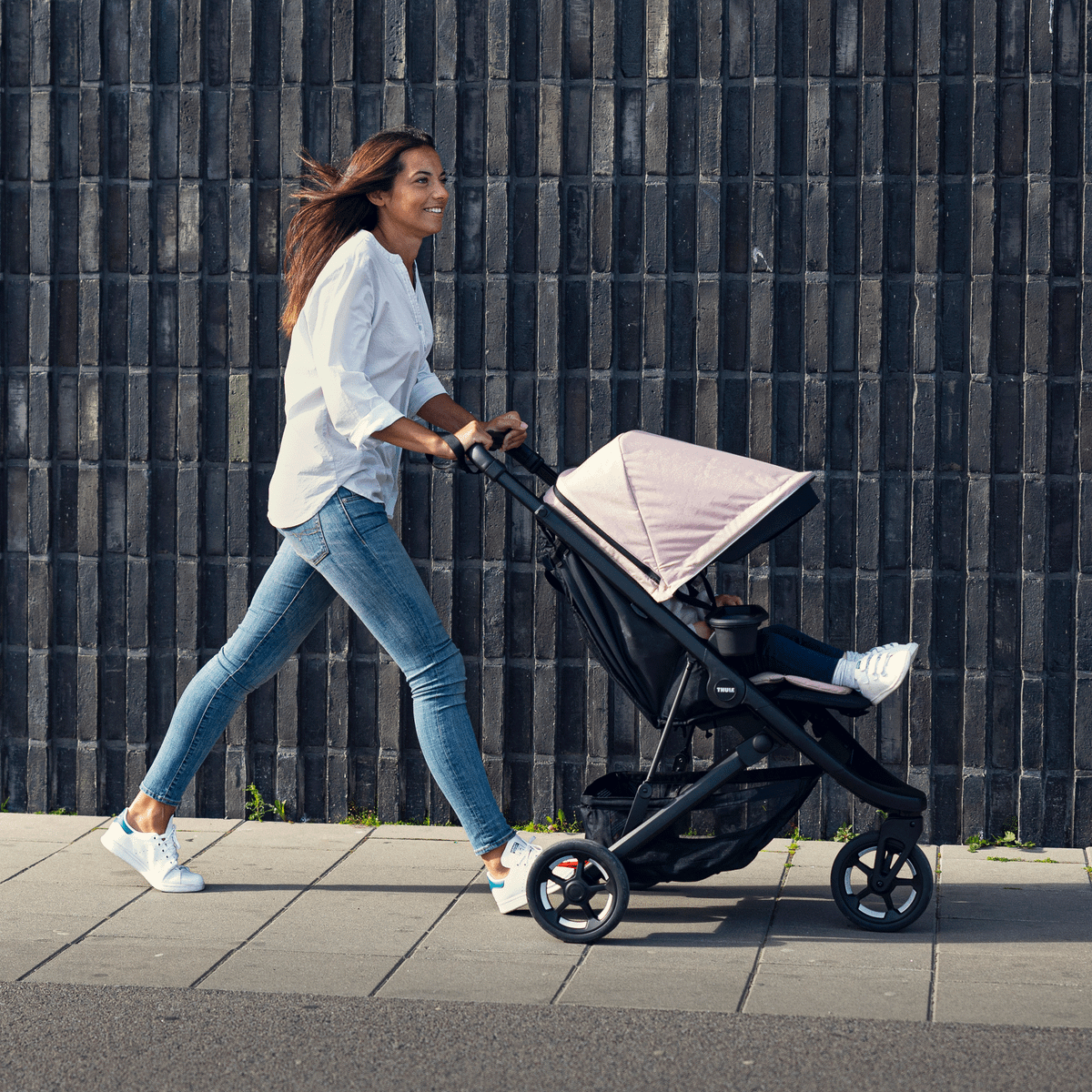 A woman pushes her pink stroller down a street and her child rests their feet on the Thule Spring Leg Rest.