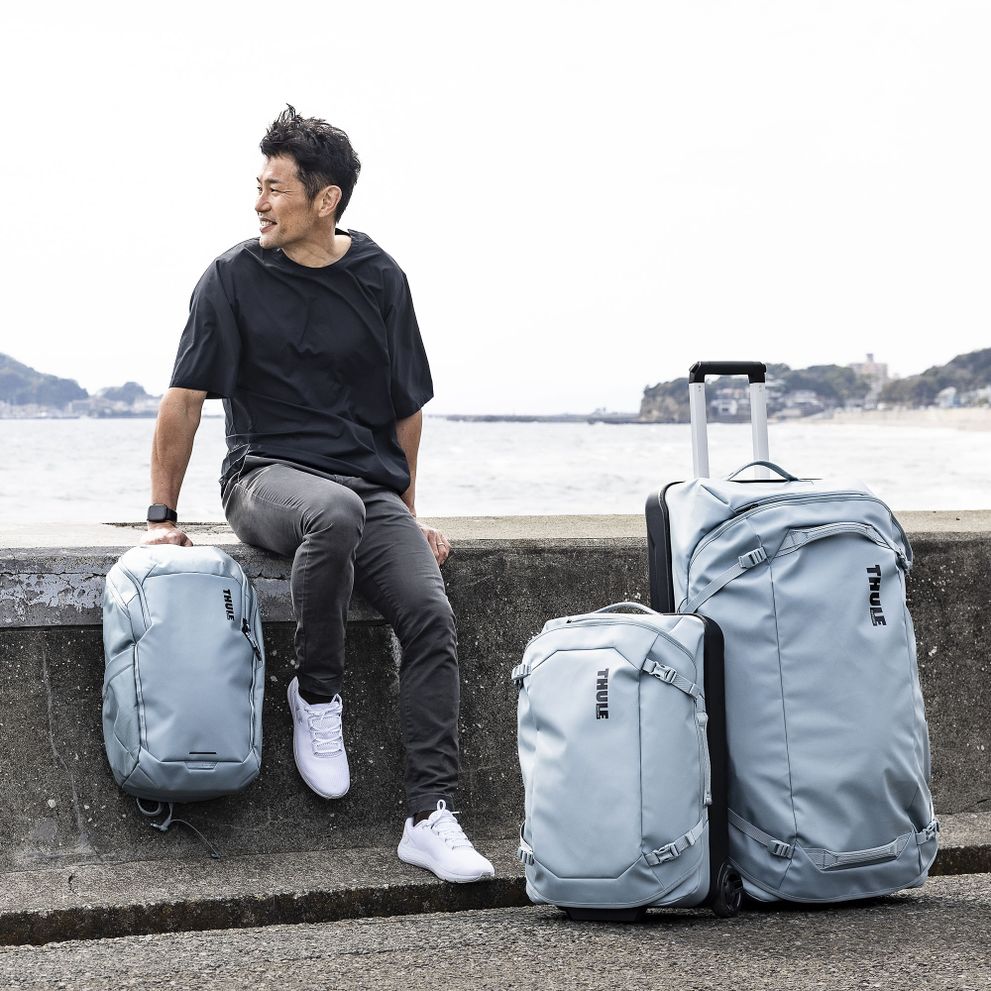 A man sits by the water with blue Thule Chasm backpack, carry-on and check-in suitcase.