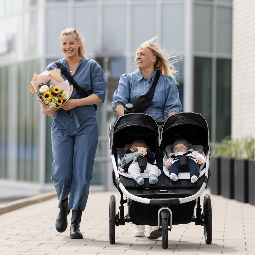 Two women wearing blue and holding flowers walk with their kids in a black Thule Urban Glide 2 double stroller.