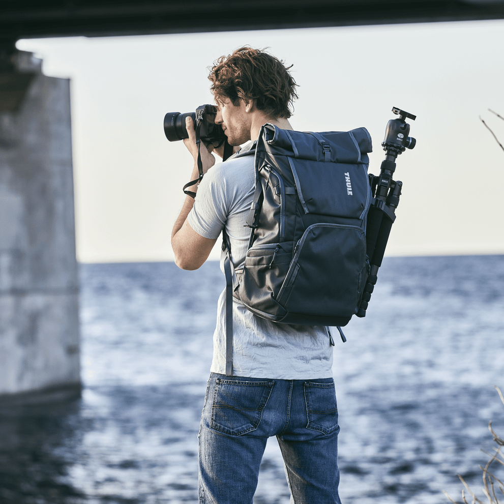A man takes a picture of the water carrying a Thule Covert DSLR camera backpack with his tripod attached.