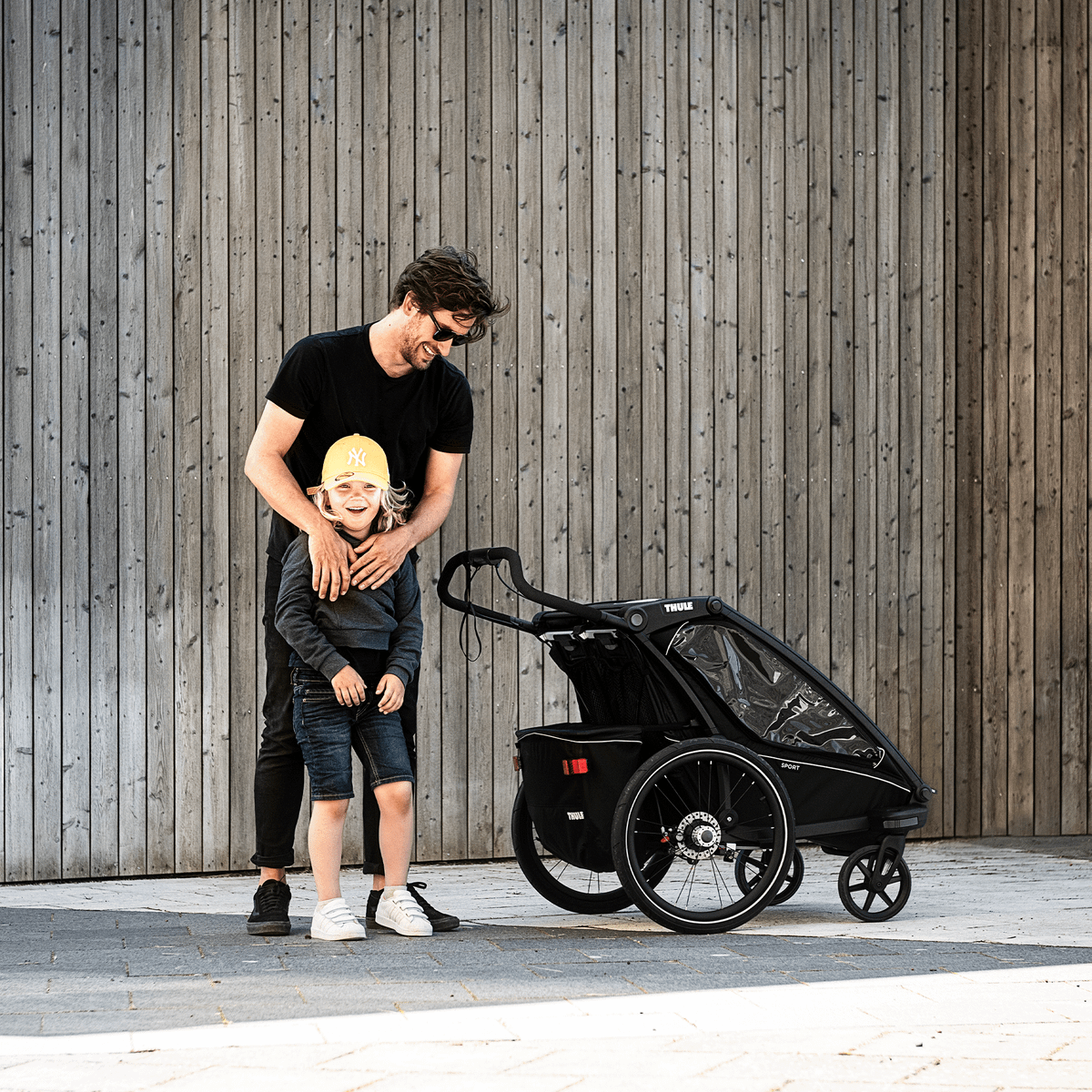A man hugs his child and stands next to a black Thule Chariot Sport child bike trailer.