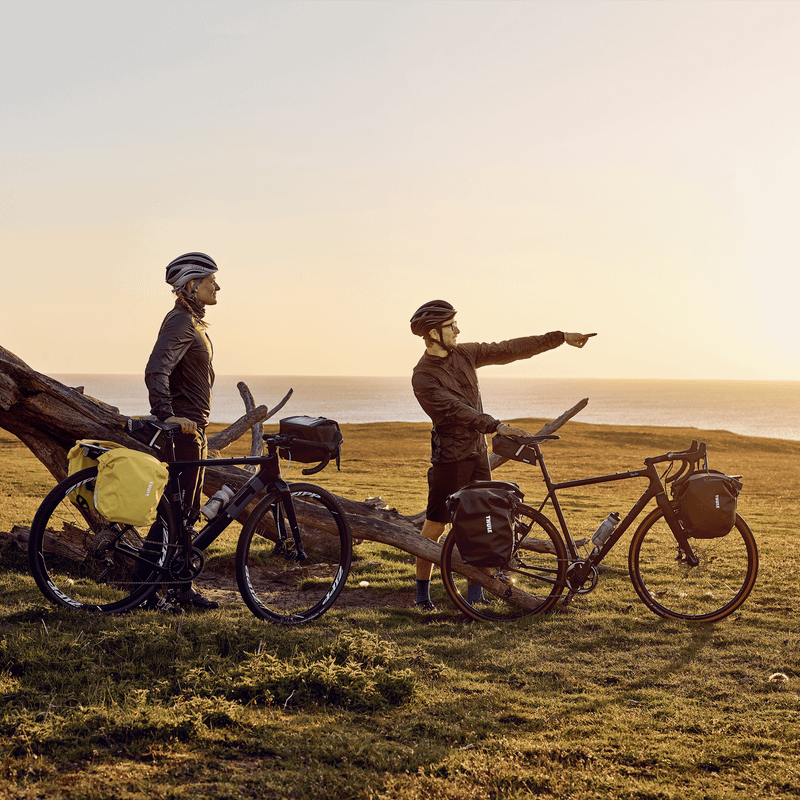 Two cyclists point at the setting sun and ocean with bike carrying 13L Thule Shield bike bags.